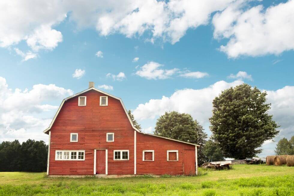 Bright red barn with white trim standing in a green grassy field under a partly cloudy blue sky, surrounded by trees and hay bales.